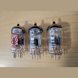 Tři elektronické vacuumové lampy s průhledným skleněným pláštěm a kovovými kontakty v dolní části. Dvě pravé lampy mají bílý nápis „TAD ECC83 Selecte…