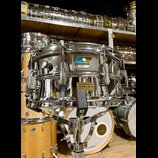 Stříbrný kovový snare drum s chromovaným povrchem, umístěný na stojanu. Na korpusu je viditelné logo "Ludwig" v modrém obdélníku a žlutá nálepka s ná…