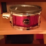Elektronický snare s fialovým třpytivým povrchem a chromovanými obručemi. Na přední straně je viditelné logo ALESIS v kruhovém medallionu a stejný ná…