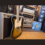 Elektrická kytara s tónem sunburst, bílým pickguardem a zlatými kovovými prvky. Kytara stojí před kytarovým zesilovačem s šedým potahem a černou mříž…