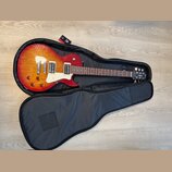 Elektrická kytara s červeně-žlutým sunburst povrchem těla a bílým pickguardem, položená v otevřeném černém softcase. Na hlavě kytary je viditelné log…