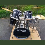 Černá bubnová sada s kovovým kováním a zlatými činely. Na basovém bubnu, tom-tamech i snare je viditelný nápis SWINGSTAR by TAMA. Na jednom z činelů…