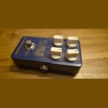 Modrý kytarový efekt s bílými ovládacími knoflíky. Na šasi jsou nápisy tc electronic, MOJOMOJO a PAUL GILBERT OVERDRIVE. Viditelné popisky u knoflíků…