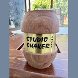 Dřevěný shaker s kovovým středovým prutem zlaté barvy. Na kovové části je černý nápis STUDIO SHAKER a svisle orientovaný text SMALL. Povrch dřeva vyk…