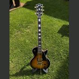 Elektrická kytara s tónem těla v barvě sunburst a černým pickguardem. Na hlavě je viditelné logo Gibson a nápis Custom. Hриф má vložky z perlemi a na…