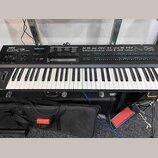 Černý digitální klavír s bílými a černými klávesami. Na levé straně je nápis YAMAHA DX7FD. Nástroj je umístěn v černém transportním kufru s kovovými…