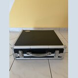 Černý transportní kufr s hliníkovými hranami a rohy. Na horní straně je nalepený oválný štítek s nápisem "DRUMCASE". Kufr má kovovou rukojeť a dva ko…