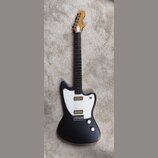 Černá elektrická kytara s bílým pickguardem a dvěma humbuckery. Hmatník je tmavého dřeva, hlava kytary je v přirozené barvě dřeva s bílým logem, kter…