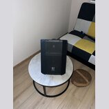 Černý reproduktor s mřížkou v horní části a logem "E" ve spodní části. Nástroj stojí na kulatém stole s mramorovým povrchem a černou kovovou nohou. P…