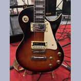 Elektrická kytara s tmelem v barvě sunburst (černá přecházející do hnědé). Má dva humbuckery s kovovými kryty, bílý pickguard a čtyři zlaté ovládací…