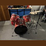Červená bicí souprava s kovovým kováním. Na tomu a snare bubnu je viditelné logo značky Pearl. Souprava obsahuje basový buben, dva tom-tomy a činely…
