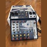 Míchací pult Soundcraft NOTEPAD-8FX v tmavě modré barvě, umístěný před svým původním bílým krabicem s nápisem Notepad 8FX. Na horní straně jsou vstup…