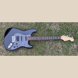 Černá elektrická kytara s šedým mramorovaným pickguardem a hnědým hmatníkem. Na hlavě je viditelné logo Fender a nápis STRATOCASTER, níže je drobný n…