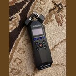 Černý digitální záznamový přístroj značky zoom. Na přední straně je displej, ovládací tlačítka a v dolní části nápis H1essential Handy Recorder. V ho…
