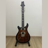 Elektrická kytara s tmavě hnědým plamenovitým povrchem těla a černou hlavou. Na hlavě je bílé logo PRS a nápis SE Custom. Hmatník obsahuje bílé vložk…