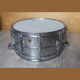 Kovový snare drum s chromovaným povrchem a bílým bubenovou skins. Na korpusu je viditelné zlaté logo ve tvaru štítu, ale text uvnitř něj není čitelný…