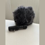 Černý mikrofon s nasazenou šedou chlupatou ochranou proti větru. Na ochraně je viditelné logo RØDE a na těle mikrofonu nápisy Videomic Me MADE IN AUS…