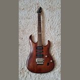 Elektrická kytara s hnědým lakovaným tělem a tmavým hnedým hmatníkem. Na hlavě je bílý nápis Cort. Nástroj má tři humbuckery, tremolo most a dvě ovlá…