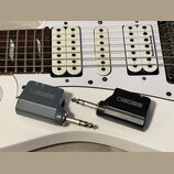 Na bílé elektrické kytáře leží dva šedé a černé adaptéry značky BOSS. Na bílém snímači kytary je viditelný nápis Seymour Duncan. Adaptéry mají kovové…
