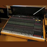 Velký míchací pult s dřevěným rámem a černým ovládacím panelem. Na pravé straně je nápis TOFT AUDIO CONSOLE MTO a logo Toft Audio Design. Panel obsah…