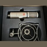 Stříbrný kondenzátorový mikrofon s mřížkou v horní části, umístěný v černé pěnové výplni. Na těle mikrofonu je červené logo s nápisem se a černý text…