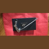 Chromovaný kytarový tremolo systém položený na černé krabici. Na krabici je bílý nápis DUESENBERG a logo ve tvaru stylizovaného písmene D s křídly. P…