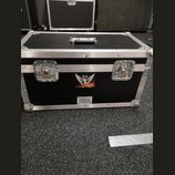Černý transportní kufr s hliníkovými hranami a kovovými rohy. Na přední straně jsou dva kovové zámky a dvě nálepky: horní s křídlatou postavou a nápi…