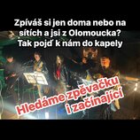 Fotografie zachicuje kapelu čtyř mužů a jedné ženy během hraní na pódiu. Jsou zde vidět elektrická kytara, akustická kytara a bicí souprava. V pozadí…