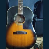 Akustická kytara s tělem v provedení sunburst (přechod od černé k žlutohnaté). Nástroj má tmavý hmatník a černý plechtr zaštitu. Uvnitř otvoru rezona…