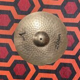 Kruhový činely z kovaného mosazi s výrazným kladivováním po celé ploše. Na povrchu jsou černé nápisy Zildjian a logo ve tvaru stylizovaného A. Na pra…