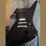 Černá elektrická kytara s ostrovním tvarem těla a hnědým hmatníkem. Nástroj má dva humbuckery, jeden ovládací knoflík a přepínač. Povrch je vysoce le…