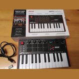 Na fotografii je kompaktní MIDI klaviatura AKAI PROFESSIONAL MPKminiplay v černé barvě s červeným okrajem, umístěná před svým původním kartonovým oba…