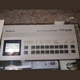 Bílý rytmický kompozér Roland RHYTHM COMPOSER TR-626. Zařízení obsahuje displej s mřížkou, několik řad ovládacích tlačítek a dva otočné ovladače ozna…
