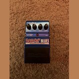 Modrý kytarový efekt s nápisy SCREAMIN' BLUES a OVERDRIVE/DISTORTION. Na pravé straně jsou čtyři ovládací knoflíky s popisky LEVEL, LOW, HIGH a GAIN…