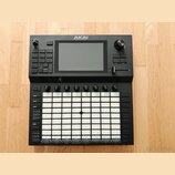 Černý MIDI kontrolér s bílou mřížkou 8x8 gumových padů. Na horní části je umístěn displej a nad ním nápis AKAI PROFESSIONAL. Zařízení obsahuje několi…
