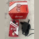 Na fotografii je červený kytarový efekt s bílými prvky, černý napájecí adaptér a červená papírová krabice. Na efektu i na krabici je nápis drop a log…