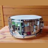 Stříbrný kovový snare drum s chromovaným povrchem. Na boku je černý čtvercový štítek s bílým nápisem MAPEX a menším textem pod ním, který je nečiteln…