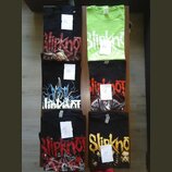 Šest triček s potisky kapely Slipknot rozložených na hnědém stole. Trička jsou v černé a neonově zelené barvě, každé má připevněný bílý lístek s ručn…