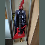 Červená elektrická kytara s ostrovním tvarem těla a hmatníkem z tmavého dřeva. Na hlavě je bílý nápis Aria. Kytara má dva humbuckery, ovládací knoflí…