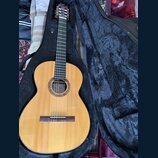 Kytara s nylonovými strunami v černém pouzdře. Na štítku uvnitř otvoru je nápis „Classical Guitar“ a pod ním podpis „Luthier Washik“. Tělo kytary je…