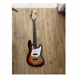 Elektrická basová kytara s tělem v barevně sunburst a bílým pickguardem. Na hlavě hředu je viditelné logo Fender. Nástroj má čtyři struny, dva snímač…