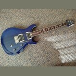 Modrá elektrická kytara s tmavým hnedým hmatníkem a bílými vložkami kolem snímačů. Na hlavě je viditelné logo značky Schecter. Kytara má dva humbucke…