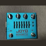 Modrý kytarový efekt s nápisy JOYO a MONOMYTH. Horní část obsahuje čtyři stříbrné otočné knoflíky s popisky VOICE, BLEND, LEVEL a DRIVE. Uprostřed je…