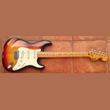 Elektrická kytara s tvarem Stratocaster, provedená v červeno-žlutém sunburst povrchu s bílým pickguardem. Na hlavě hředu je vidět logo Greco a nápis…