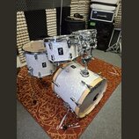 Bílá perleťová bubnová sada značky SONOR. Sestává z basového bubnu, tomů a malého bubnu s kovovým hardwarem. Na jednom z tomů je viditelný nápis AC2…