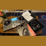 Na fotografii je černý DJ 컨트롤er značky Numark s modrým displejem a dvěma jogovými koly. Vedle něj leží krabice a černé sluchátka s nápisem Pioneer DJ…