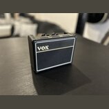Malý kytarový zesilovač s černým povrchem z texturovaného materiálu a bílými lemovými pruhy. Na přední straně je umístěno zlaté logo VOX. Horní panel…