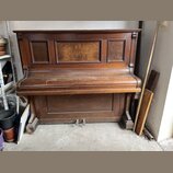 Hnědé dřevěné piano s výraznou masivní konstrukcí a dekorativními panely. Povrch je pokryt prachem a vykazuje známky opotřebení v podobě škrábanců. N…