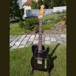 Elektrická kytara s černým bodies, hnědým hnedem na okrajích a tmavým hmatníkem. Na hlavě je viditelné logo Fender Telecaster. Kytara má černý pickgu…