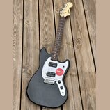 Černá elektrická kytara s bílým pickguardem a hnědým hmatníkem. Na hlavě je viditelné logo Squier. Kytara má dva snímače a na těle je nalepená červen…